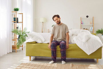 Man is trying to wake up from sleep on a very early Monday morning. Sleepy, tired man in a T shirt and pajama pants is sitting with closed eyes on the couch at home