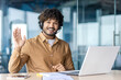 © Liubomir - Portrait of a smiling Indian young man wearing a suit sitting in the office at a desk with a laptop, looking and waving at the camera.