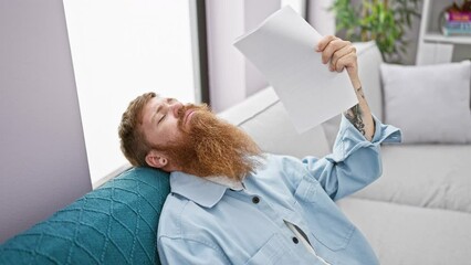 Poster - Redhead young man, suffering in hot living room, sitting on sofa, making air with documents to cool down