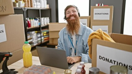 Sticker - Smiling young redhead man enjoying volunteering at a charity center, sitting at a table with his laptop and headphones on