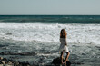 Kobieta w białej sukience stoi tyłem na kamienistej plaży i patrzy w ocean