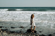 Kobieta w białej sukience stoi tyłem na kamienistej plaży i patrzy w ocean