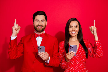 Poster - Portrait of two cheerful elegant people toothy smile hold smart phone direct finger up empty space isolated on red color background