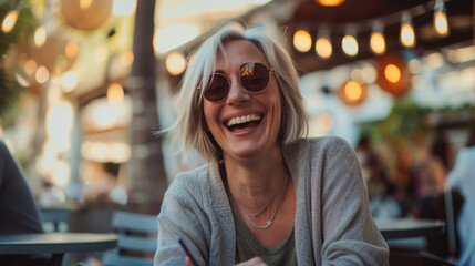 Wall Mural - A cheerful young woman wearing round glasses and a casual sweater is laughing heartily while sitting in a caf?.