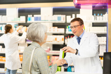 Polite middle-aged male pharmacist consulting old woman costumer about care product in box in chemist's shop