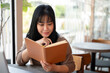 © bongkarn - A happy Asian female college student is diligently reading a book in a cafe co-working space.