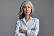 © The Picture Show - Stylish confident adult 50 years old Asian female psychologist standing arms crossed looking at camera at gray background. Portrait of sophisticated grey hair woman advertising products, Generative AI