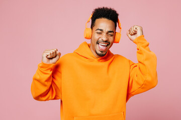 Wall Mural - Young man of African American ethnicity wears yellow hoody casual clothes listen to music in headphones raise up hands dance isolated on plain pastel light pink background studio. Lifestyle concept.