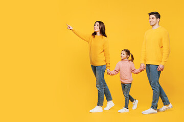 Full body side view young parent mom dad with child kid girl 7-8 year old wear pink sweater casual clothes hold hands walk go point finger aside isolated on plain yellow background Family day concept