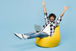 © ViDi Studio - Full body young Indian IT man he wears shirt white t-shirt casual clothes sit in bag chair hold use work on laptop pc computer raise up hands isolated on plain blue cyan background. Lifestyle concept.