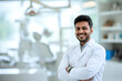 © boxstock production - Handsome Indian male dentist smiling, standing with folded hands inside blurry modern clinic. Dental care concept
