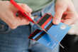 © Studio Marmellata - A person in a green sweater is seen cutting a stripe-patterned credit card with red scissors. The act signifies the intention to reduce debt or prevent overspending