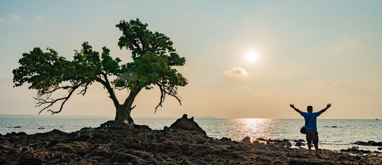 Wall Mural - Silhouette, sunset view with seaside trees and man standing, man standing with his hands up, happily watching the sunset.