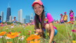 A smiling girl in a pink shirt kneels among colorful flowers with a city skyline in the background.