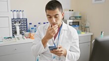 Shocked Young Hispanic Guy In Lab Coat Covers Mouth In Surprise, Scared After Seeing Smartphone Results