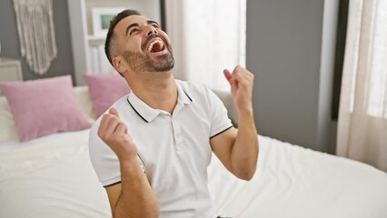 Poster - Joyful young hispanic man celebrating a triumphant win, excitedly screaming 'yes' while sitting on the bed in his bedroom.