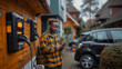 © AS Photo Family - African american man using mobile phone while standing in front of house and charging his electric car.