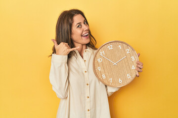 Wall Mural - Woman holding clock on yellow backdrop showing a mobile phone call gesture with fingers.