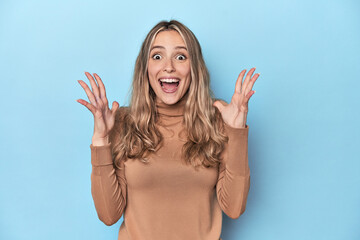 Wall Mural - Blonde young caucasian woman in blue studio celebrating a victory or success, he is surprised and shocked.