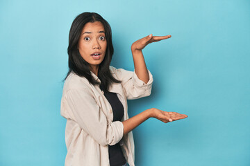 Wall Mural - Filipina young woman on blue studio shocked and amazed holding a copy space between hands.