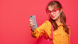 Stylish student with a backpack dressed in yellow saying something , eyes to the viewer, holding up a cell phone with a flirty playful look - Red backdrop copy space