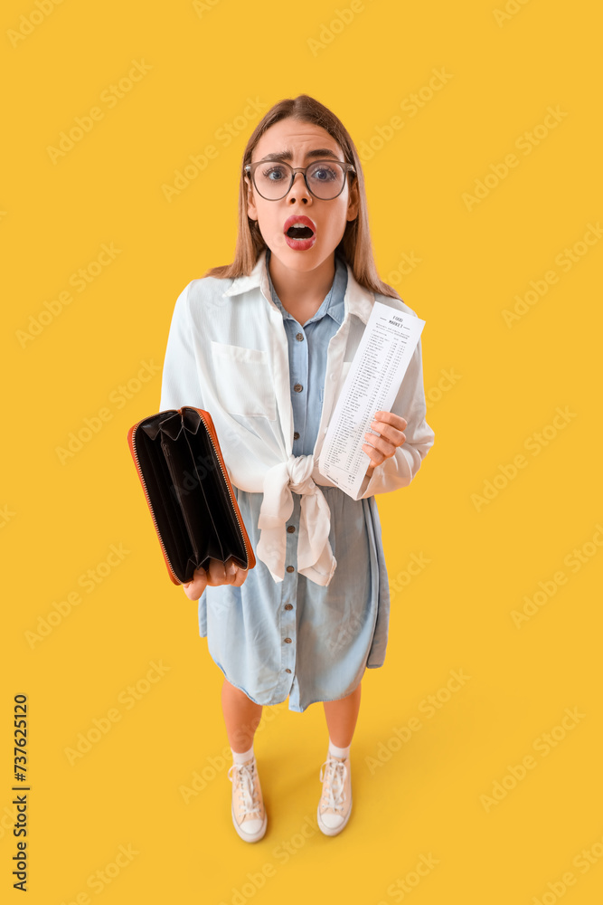 Shocked young woman with empty wallet and store receipt on yellow background. Price rise concept