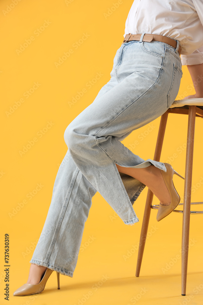 Stylish young woman in jeans with stool on yellow background
