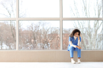 Poster - Tired female African-American medical intern sitting on windowsill in clinic