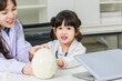 Asian woman doctor physiotherapist explains human fake skeleton skull model on a table to little cute children girl at laboratory study room. Education anatomical human concept learning for kids.
