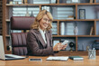 © Liubomir - Cheerful mature businesswoman using a smartphone in a well-appointed office, embodying professionalism and confidence.