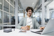 © Liubomir - Smiling young businessman with curly hair and headphones working at a laptop in a well-lit office environment.