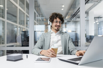 Smiling young businessman with curly hair and headphones working at a laptop in a well-lit office environment.