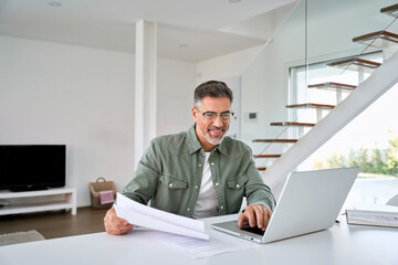 Wall Mural - Happy middle aged mature man entrepreneur wearing eyeglasses looking at laptop holding paper bill checking financial invoice or tax document making online payments working at home.