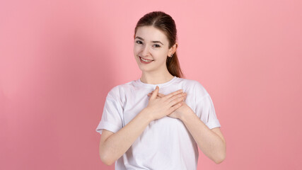 Wall Mural - Portrait of a touched attractive Caucasian young brunette woman in a casual white t-shirt, touches the heart, the girl is grateful and says thank you, on a pink studio background.