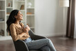 © Home-stock - Young Indian lady sitting in comfy armchair, drinking coffee, taking break or enjoying morning hot beverage at home, free space