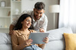 © Home-stock - Happy indian spouses using digital tablet, surfing internet or shopping online while resting on sofa at home, free space. Hindu man and woman relaxing with modern gadget