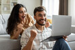 © Home-stock - Happy hindu spouses celebrate victory using laptop, shake fists in joy, embodying success and happiness, browsing online on computer at home