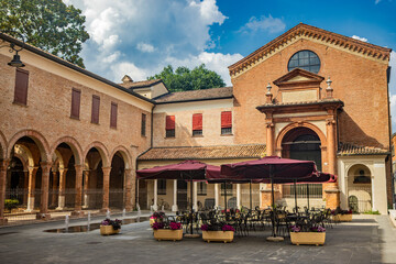 Wall Mural - Ferrara, Emilia Romagna, Italy. A characteristic view of the city, with a church, a portico, the tables of a bar sheltered by umbrellas. UNESCO World Heritage Site.