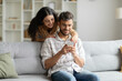 © Home-stock - Young hindu couple sharing social media on cellphone, happy indian spouses sitting on sofa, using smartphone gadget at home interior, woman embracing husband from back