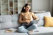© Home-stock - Indian lady wearing headphones and using laptop, enjoying digital leisure and e-learning, sitting on sofa at home interior, copy space