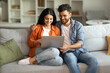 © Home-stock - Loving indian spouses using laptop together, sitting on sofa in cozy home interior, man hugging his wife and looking at computer screen, choosing movie