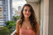 © Sascha - Smiling young brazilian woman with wavy hair on a balcony with a cityscape backdrop, wearing a salmon tank top.