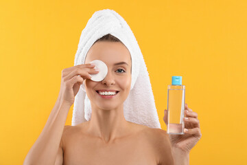 Wall Mural - Smiling woman removing makeup with cotton pad and holding bottle on yellow background