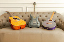 Three Guitars Set On A Brown Leather Sofa In The Living Room. A Black Electric Guitar, A Brown Acoustic And A Purple Classical Guitar. Indoors At Home With Moldings On A Grey Wall In The Background. 