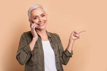 Poster - Portrait of smiling mature age business woman talk phone call with partner point finger copyspace proposition over beige color background