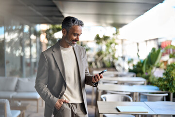 Wall Mural - Happy smiling middle aged business man, rich mature professional businessman entrepreneur standing in outdoor cafe holding smartphone using mobile cell phone digital tech. Authentic candid photo.