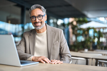 Wall Mural - Happy confident mature professional business man hybrid working looking at camera. Smiling middle aged businessman wearing glasses sitting at outdoor office table using laptop. Portrait. Copy space.