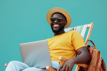 Joyful emotional African American man in summer hat sunglasses with laptop and holiday travel bag sit on beach chair by blank copy space wall