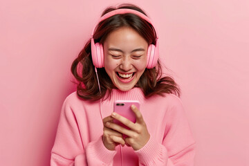 Wall Mural - Happy cheerful joyful positive young Japanese woman on pastel studio background uses mobile phone and noise cancellation headphones head set, has good time, enjoys her favorite tracks