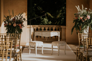 altar de ceremonia de boda. Mesa y dos sillas pequeñas. dos centros florales a los lados. Hay varias sillas de color dorado.